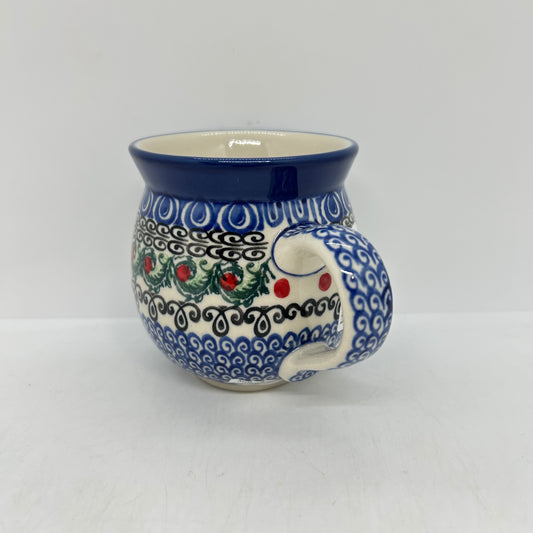 Side profile view of a Ceramika Artystyczna Polish pottery bubble mug, focusing on the curved handle decorated in a blue wave pattern alongside the green leaves and red berry motifs.