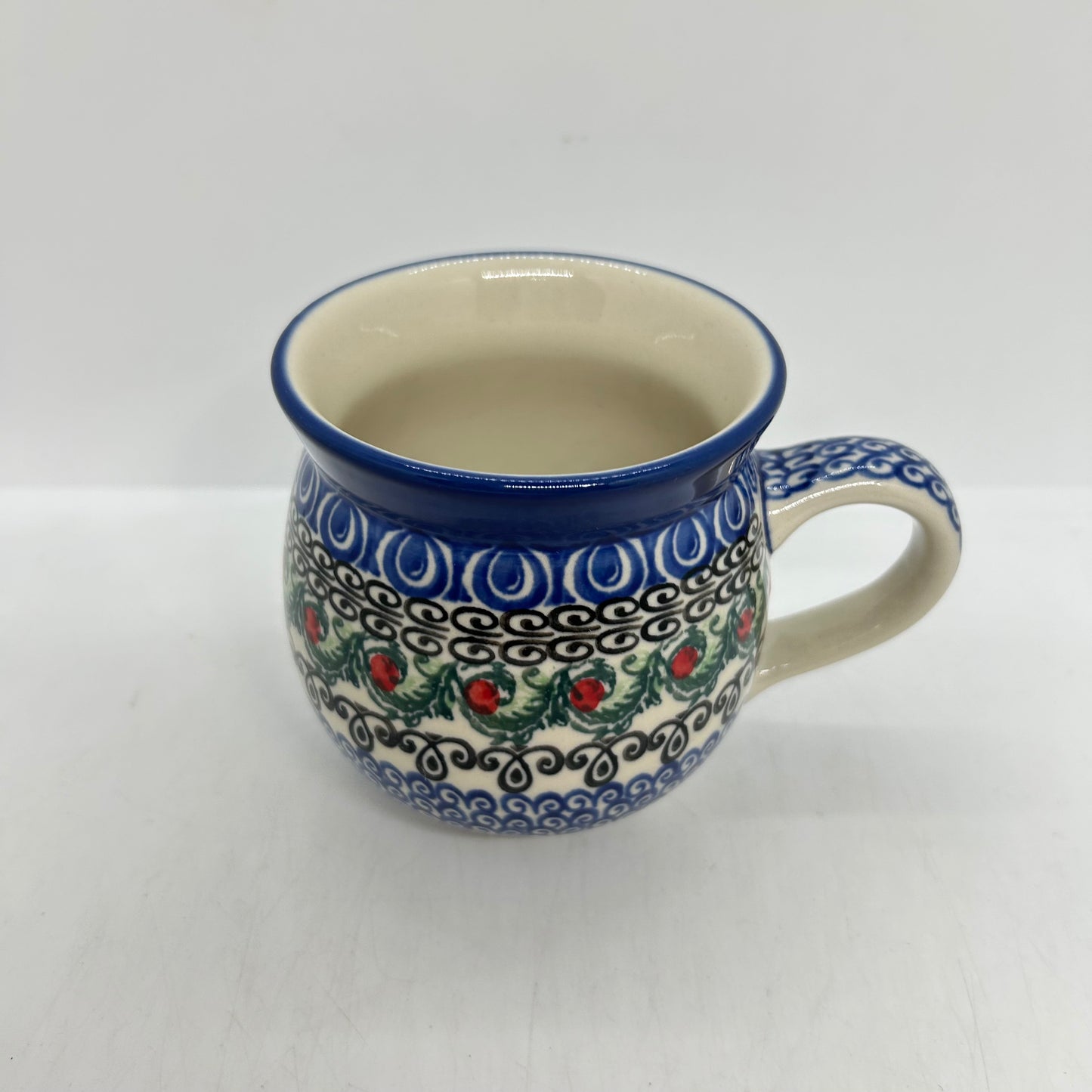Elevated top-down view of a Ceramika Artystyczna Polish pottery bubble mug, highlighting the clean off-white interior, flared blue rim, and the exterior green leaves and red berry pattern.