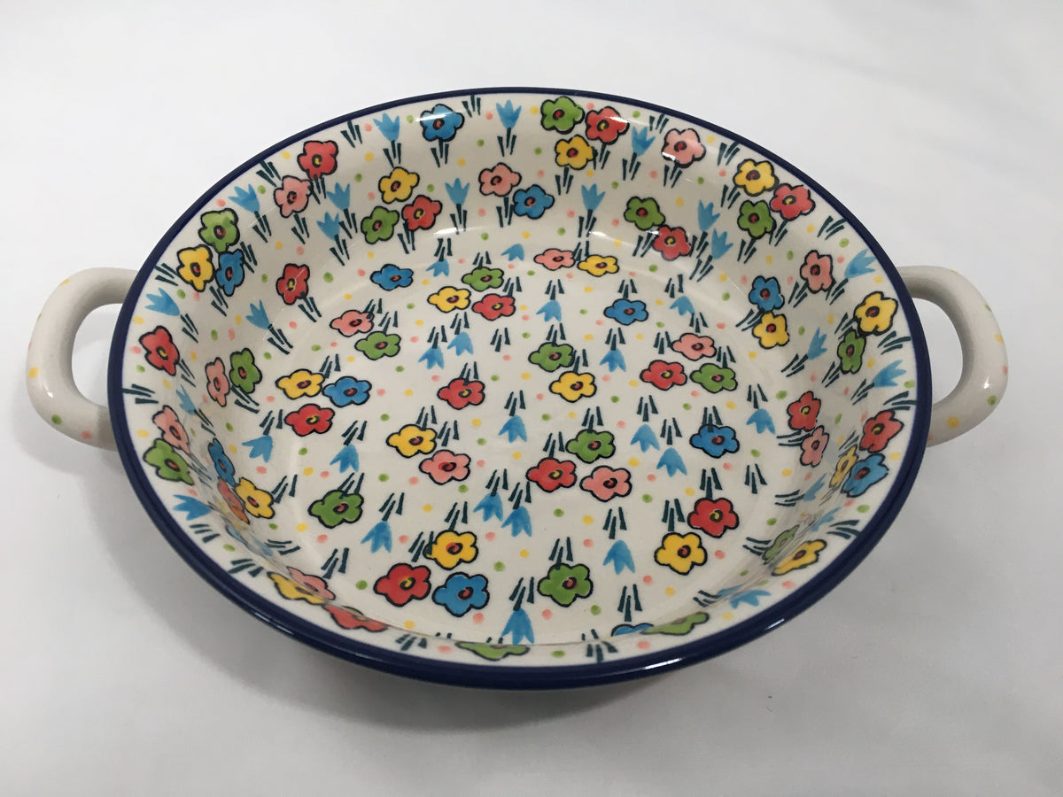 Elevated angled view of a Ceramika Artystyczna Polish pottery round baker featuring dual side loop handles, a dark blue rim, and hand-painted multi-colored spring flowers covering the interior and exterior.