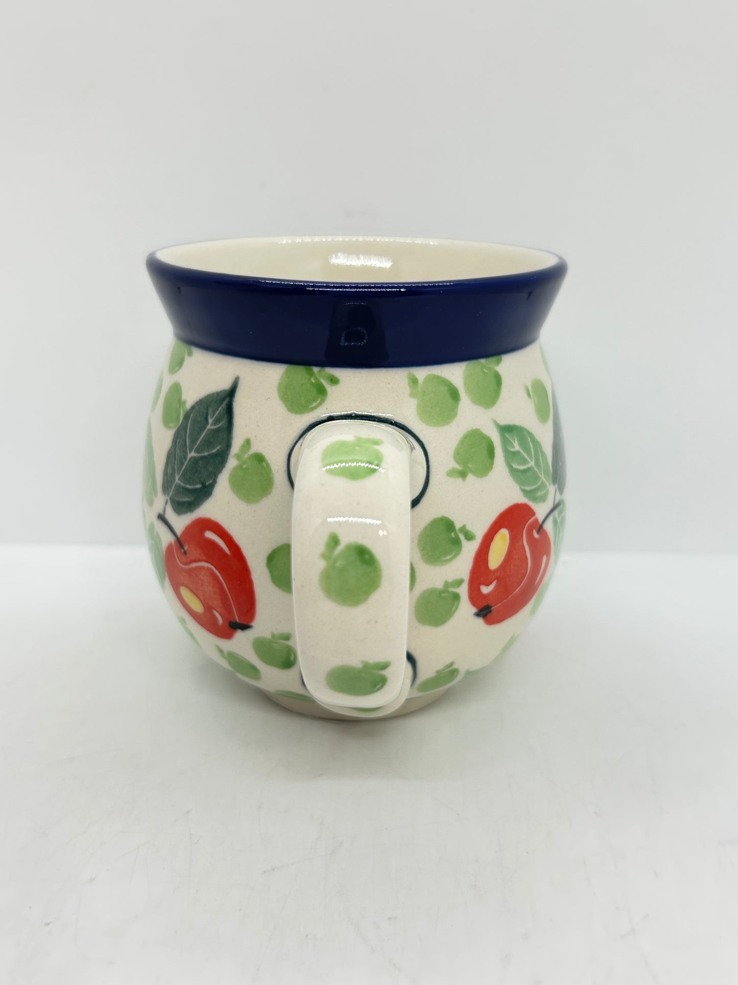 Straight-on view focusing on the patterned handle of a Ceramika Artystyczna Polish pottery bubble mug, flanked by red and green apples, green leaves, and a solid dark blue rim.