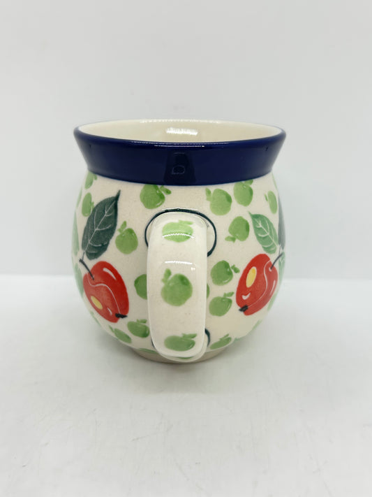 Straight-on view focusing on the patterned handle of a Ceramika Artystyczna Polish pottery bubble mug, flanked by red and green apples, green leaves, and a solid dark blue rim.