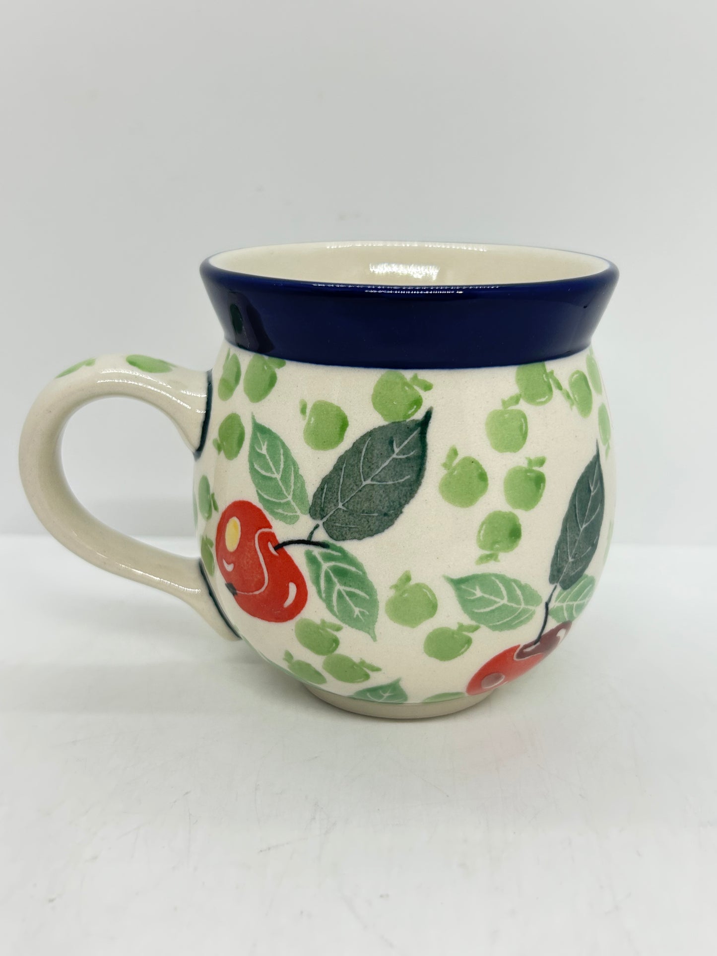 Straight-on side profile of a Ceramika Artystyczna Polish pottery bubble mug with the handle on the left, featuring hand-painted red and green apples, green leaves, and a solid dark blue rim.