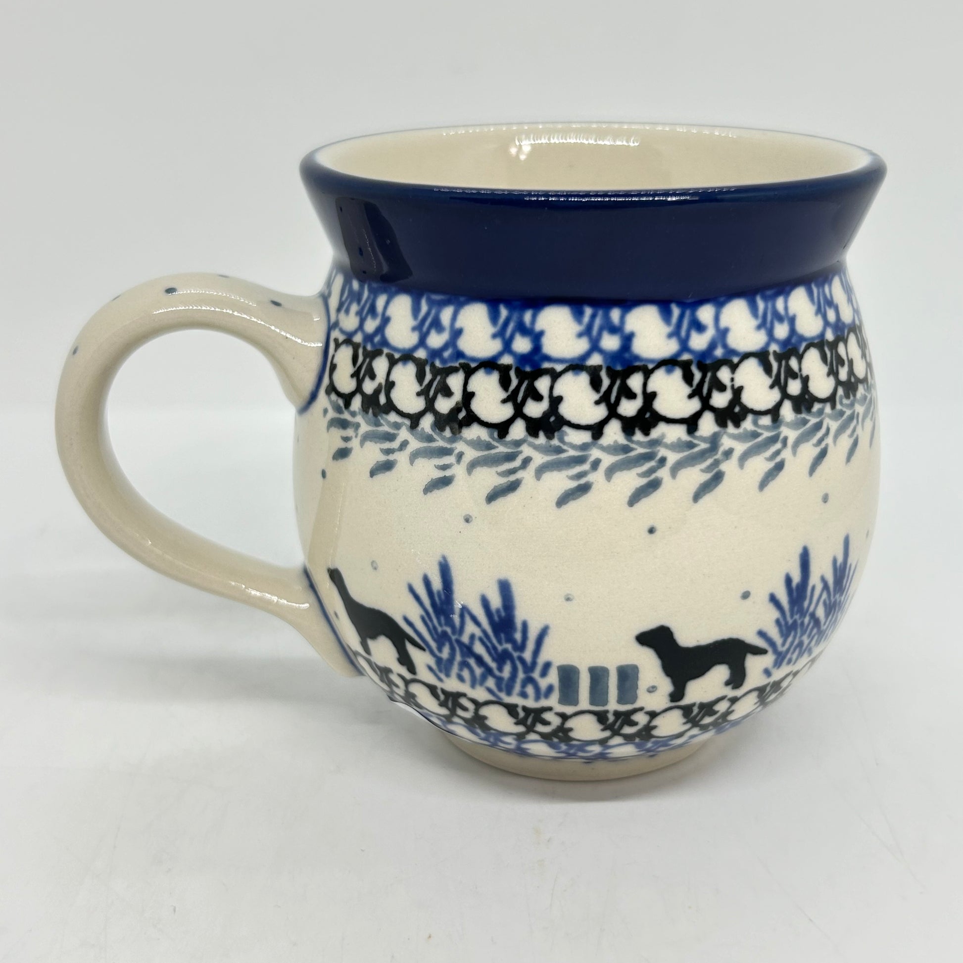 Straight-on side profile of a Ceramika Artystyczna Polish pottery bubble mug with the handle on the left, featuring black dog silhouettes, blue plants, layered borders, and a solid dark blue rim.