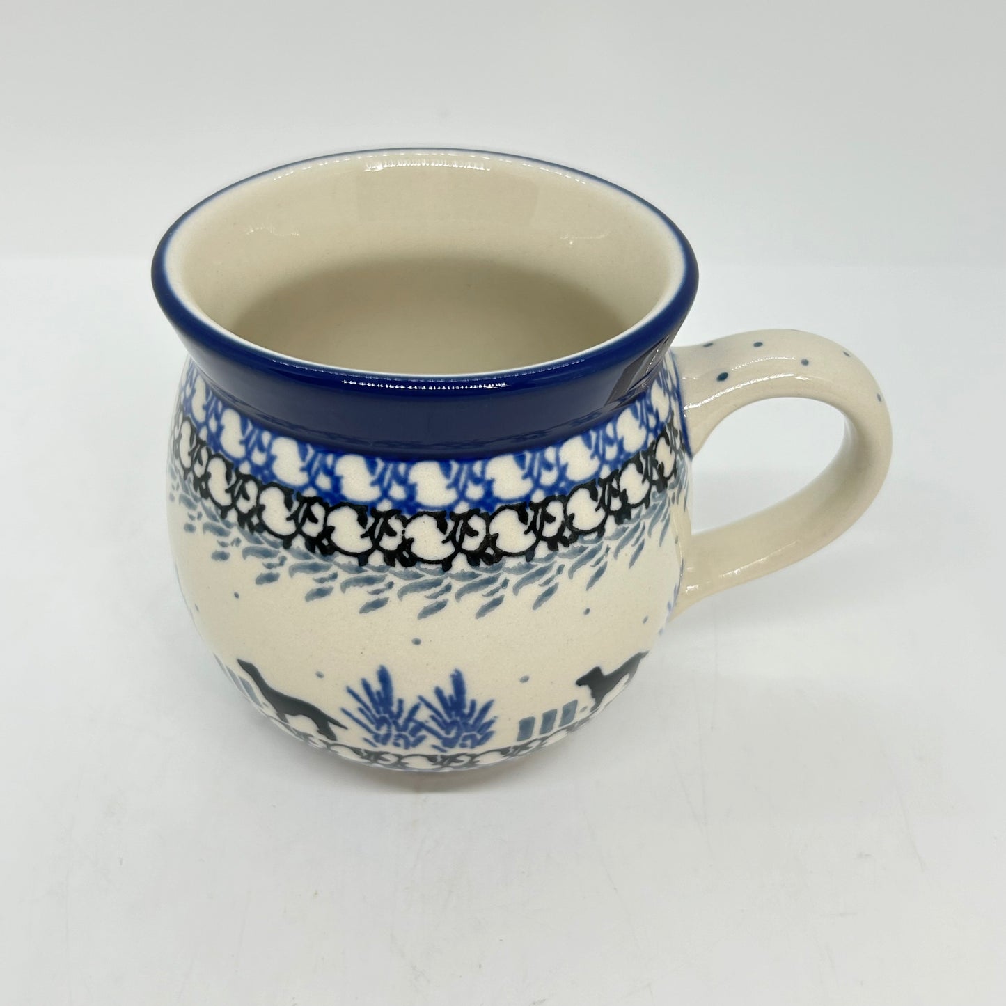 Elevated three-quarter view of a Ceramika Artystyczna Polish pottery bubble mug featuring hand-painted black dog silhouettes, complex layered borders, a dotted right handle, and a smooth off-white interior.