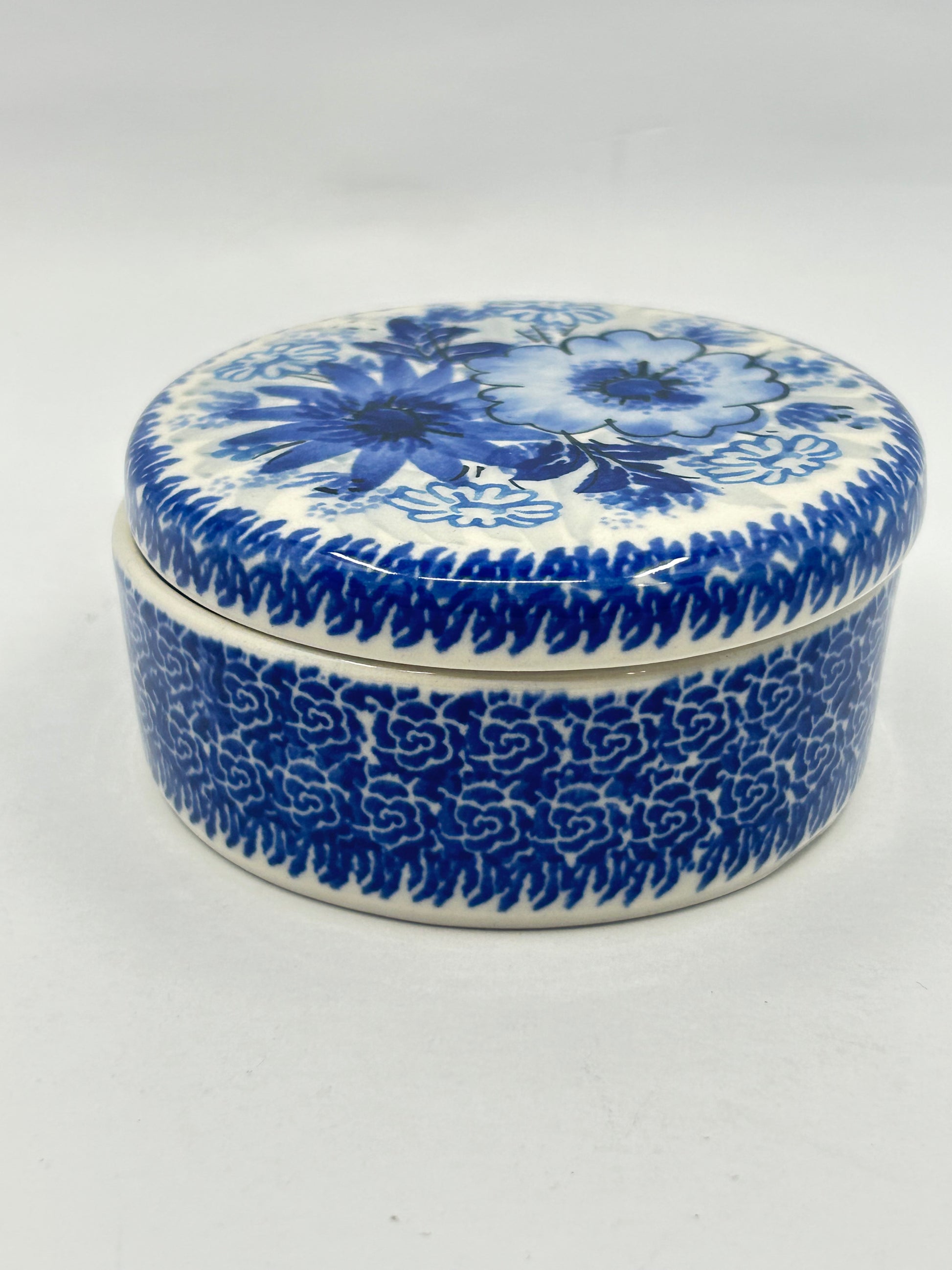 Elevated side view of a round Ceramika Artystyczna Polish pottery trinket box featuring a blue-and-white floral lid and a dense, intricate repeating blue pattern on the base.