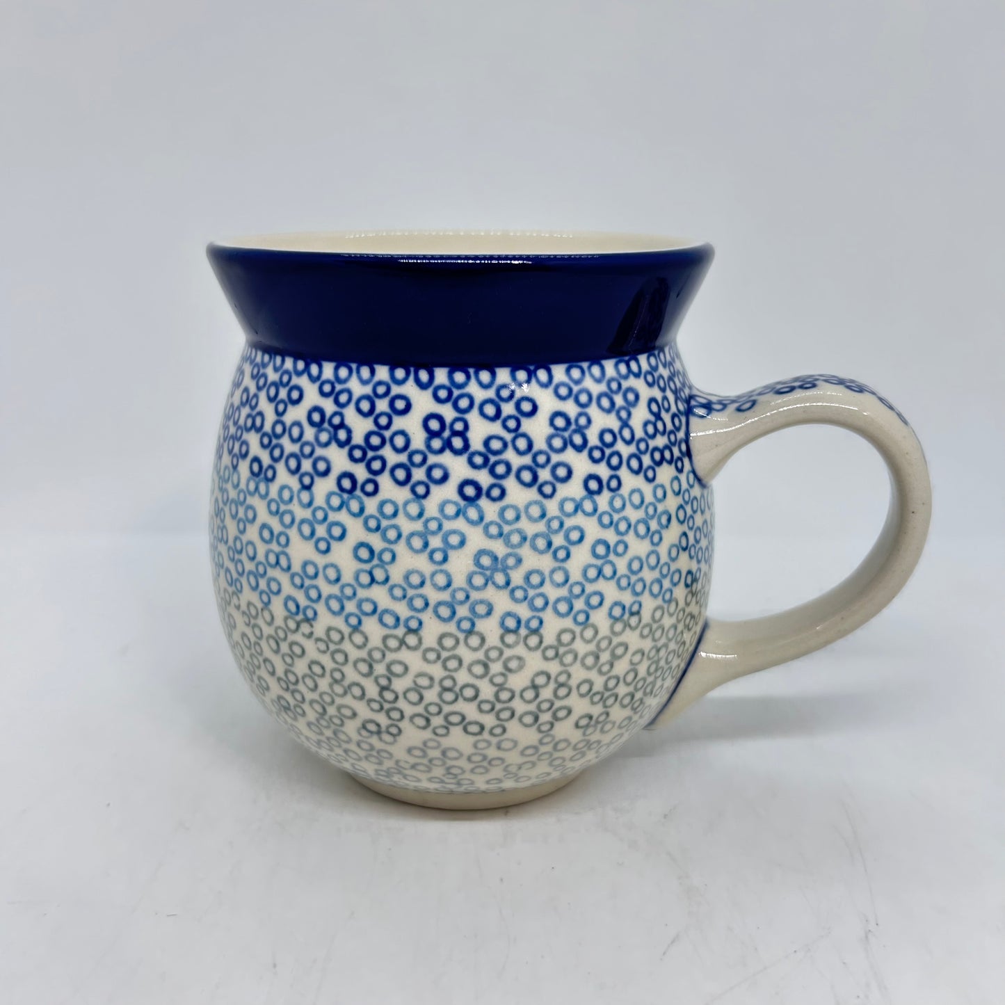 Elevated view of a Ceramika Artystyczna Polish pottery bubble mug, showing a clean cream interior, a solid dark blue rim, a dotted handle, and an exterior featuring a dense gradient of dark blue, light blue, and grey stamped circles.