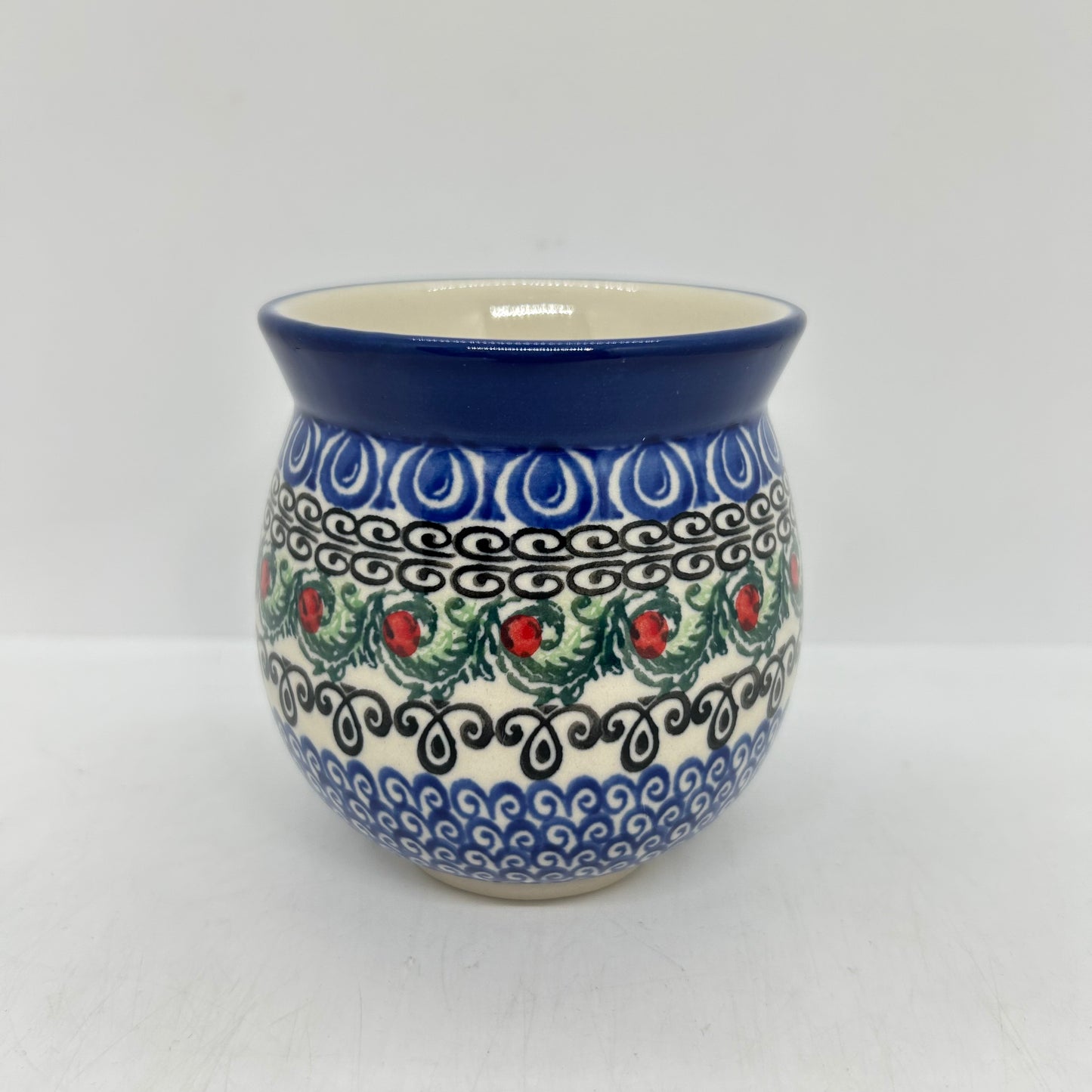 Straight-on view of the back of a Ceramika Artystyczna Polish pottery bubble mug, showing the continuous horizontal bands of blue wave motifs, black scrollwork, and green leaves with red berries without the handle visible.