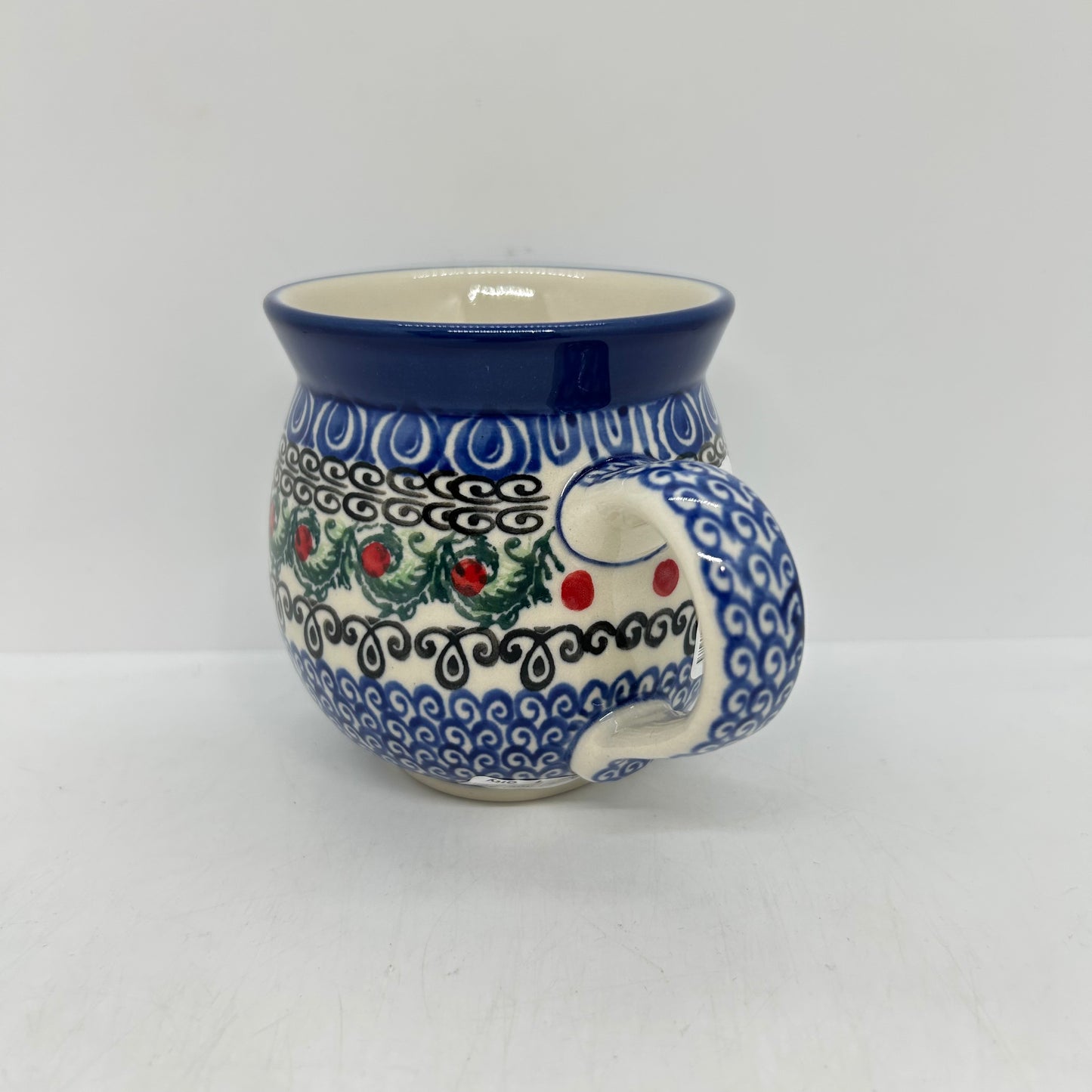 Side profile view of a Ceramika Artystyczna Polish pottery bubble mug, focusing on the curved handle decorated in a blue wave pattern alongside the green leaves and red berry motifs.