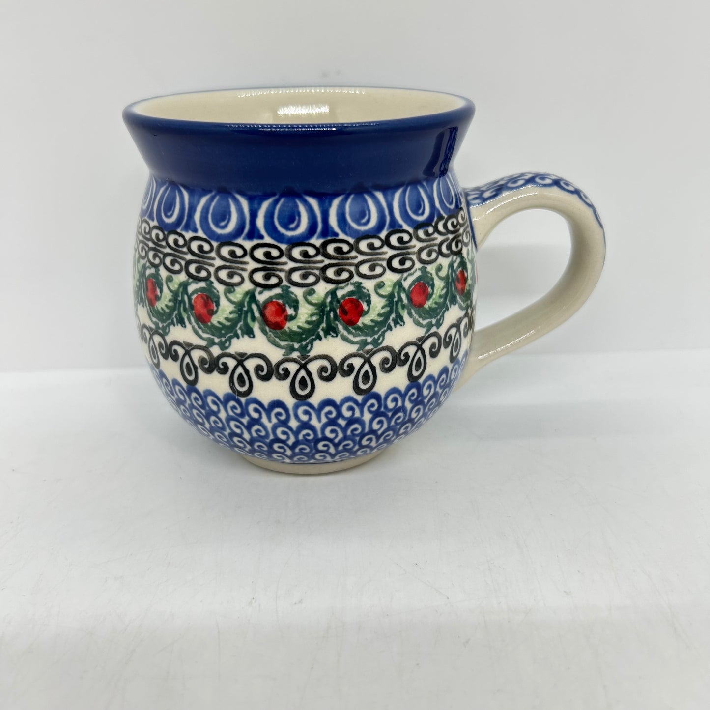 Angled front view of a Ceramika Artystyczna Polish pottery bubble mug featuring horizontal bands of blue, black scrollwork, and central green leaves with red berries.