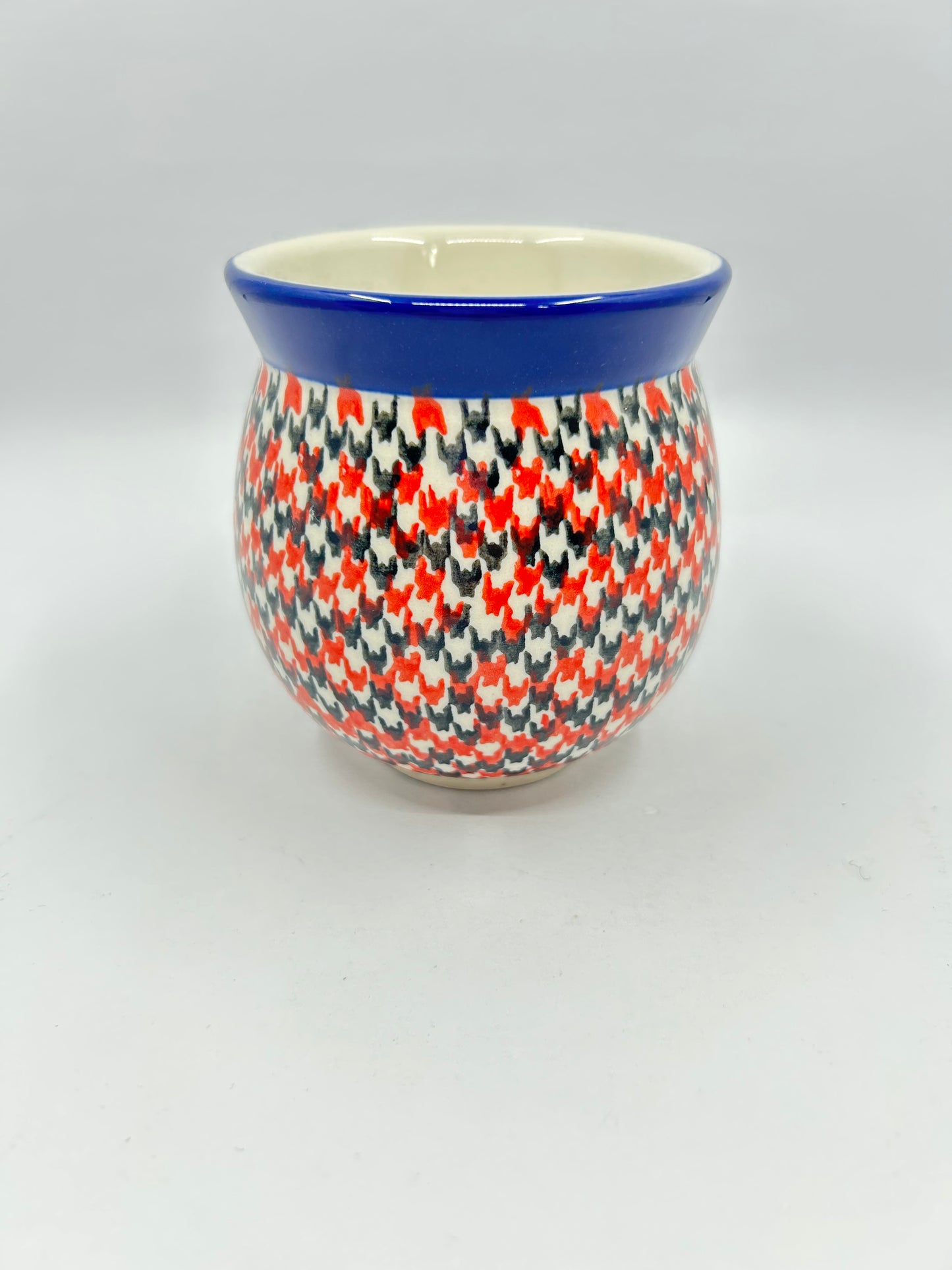 Straight-on view of the back of a Ceramika Artystyczna Polish pottery bubble mug, showing a continuous dense red, orange, and dark grey geometric pattern and a solid dark blue rim with no handle visible.