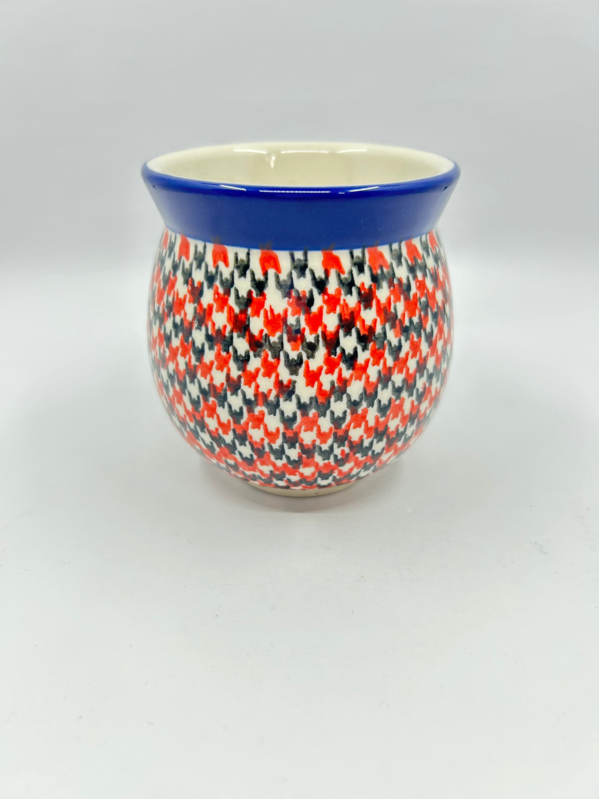 Straight-on view of the back of a Ceramika Artystyczna Polish pottery bubble mug, showing a continuous dense red, orange, and dark grey geometric pattern and a solid dark blue rim with no handle visible.
