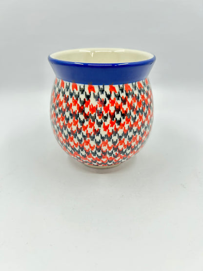 Straight-on view of the back of a Ceramika Artystyczna Polish pottery bubble mug, showing a continuous dense red, orange, and dark grey geometric pattern and a solid dark blue rim with no handle visible.