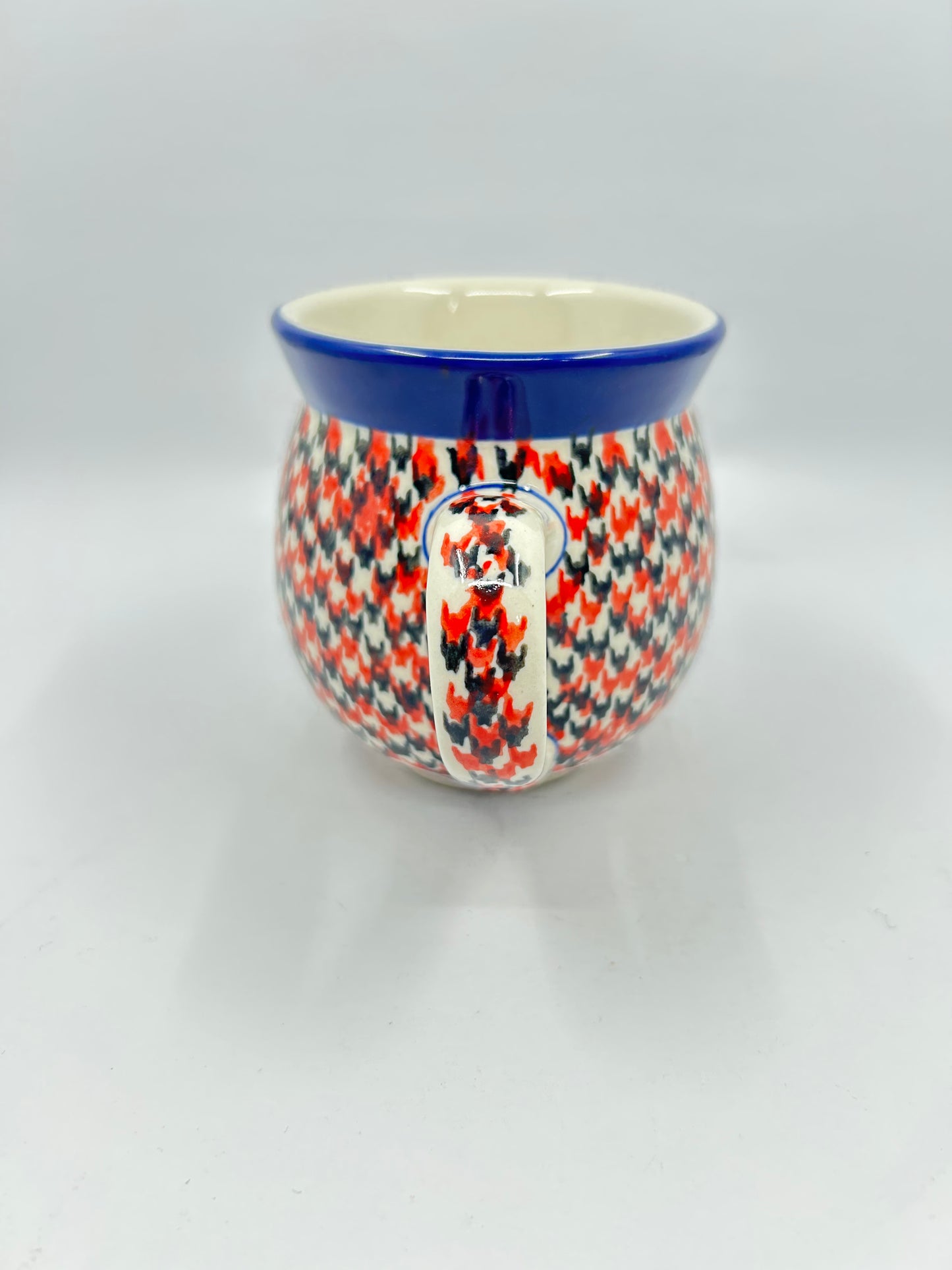 Straight-on view focusing on the patterned handle of a Ceramika Artystyczna Polish pottery bubble mug, flanked by a dense red, orange, and dark grey geometric pattern and a solid dark blue rim.