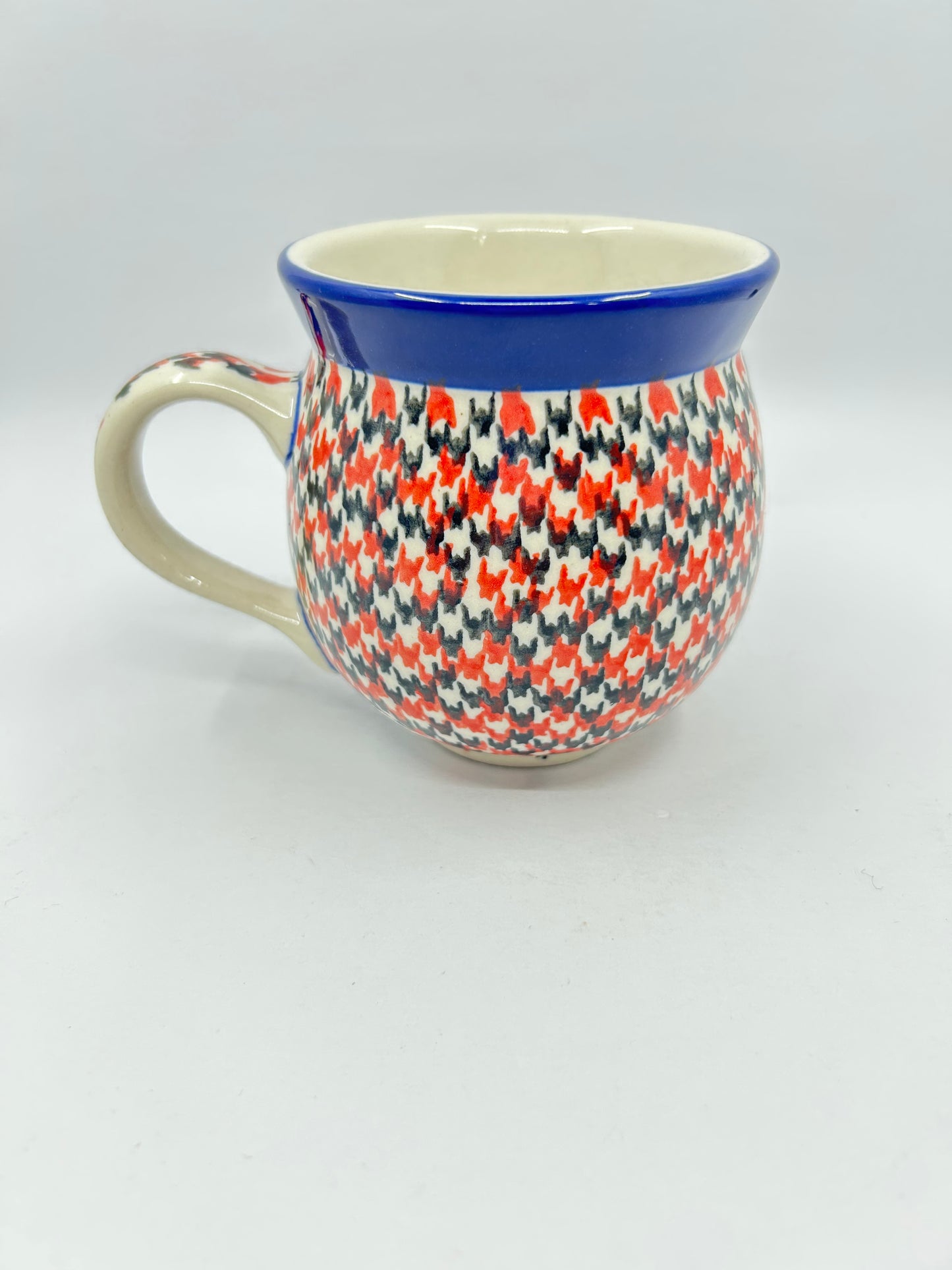Straight-on side profile of a Ceramika Artystyczna Polish pottery bubble mug with the handle on the left, completely covered in a dense red, orange, and dark grey geometric pattern with a solid dark blue rim.