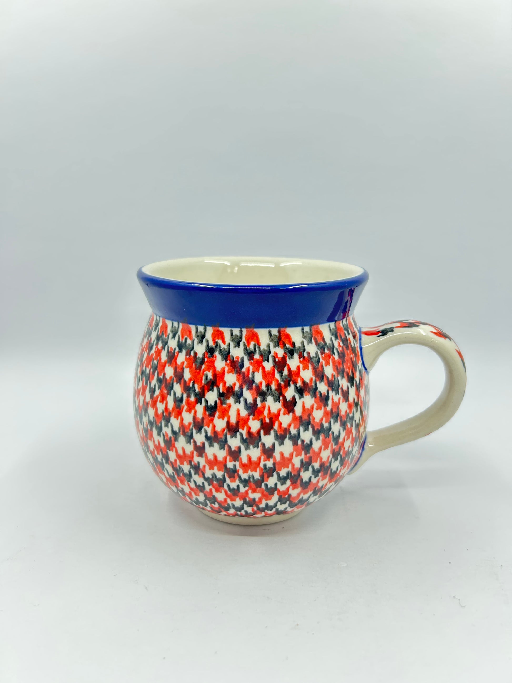 Straight-on side profile of a Ceramika Artystyczna Polish pottery bubble mug with the handle on the right, completely covered in a dense red, orange, and dark grey geometric pattern with a solid dark blue rim.