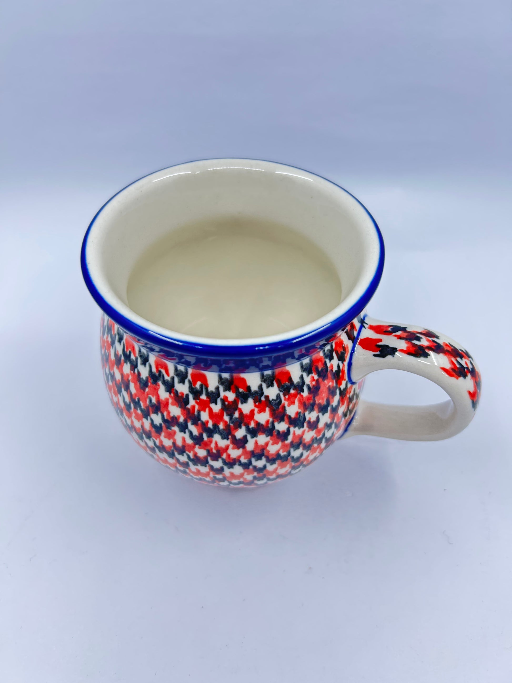 Elevated three-quarter view of a Ceramika Artystyczna Polish pottery bubble mug featuring a dense red, orange, and dark grey geometric pattern reaching a thin blue rim, a patterned right handle, and a smooth off-white interior.