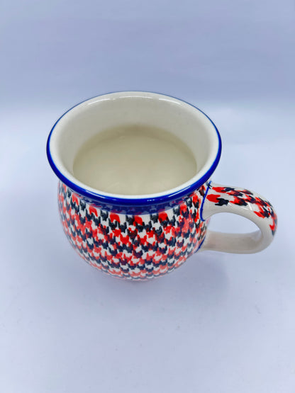 Elevated three-quarter view of a Ceramika Artystyczna Polish pottery bubble mug featuring a dense red, orange, and dark grey geometric pattern reaching a thin blue rim, a patterned right handle, and a smooth off-white interior.