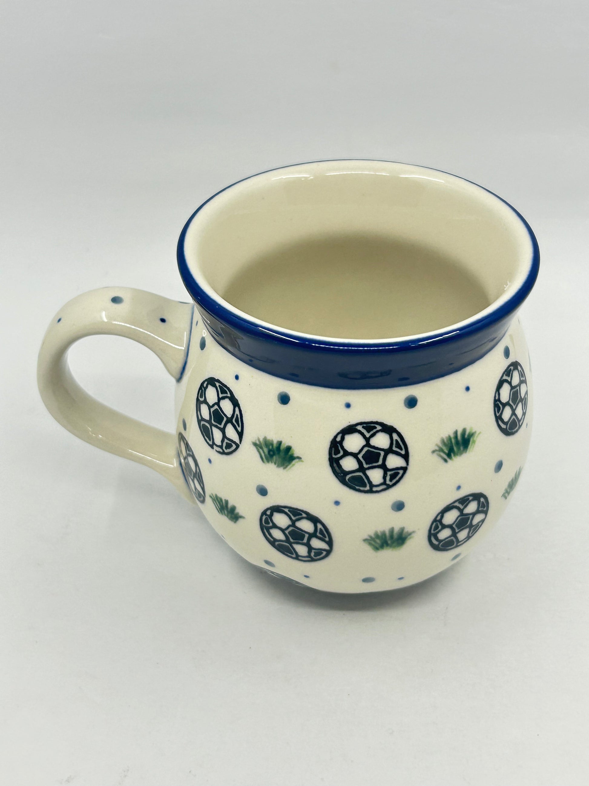 Elevated three-quarter view of a Ceramika Artystyczna Polish pottery bubble mug with the handle on the left, displaying a hand-painted soccer ball pattern, dark blue rim, and smooth off-white interior.