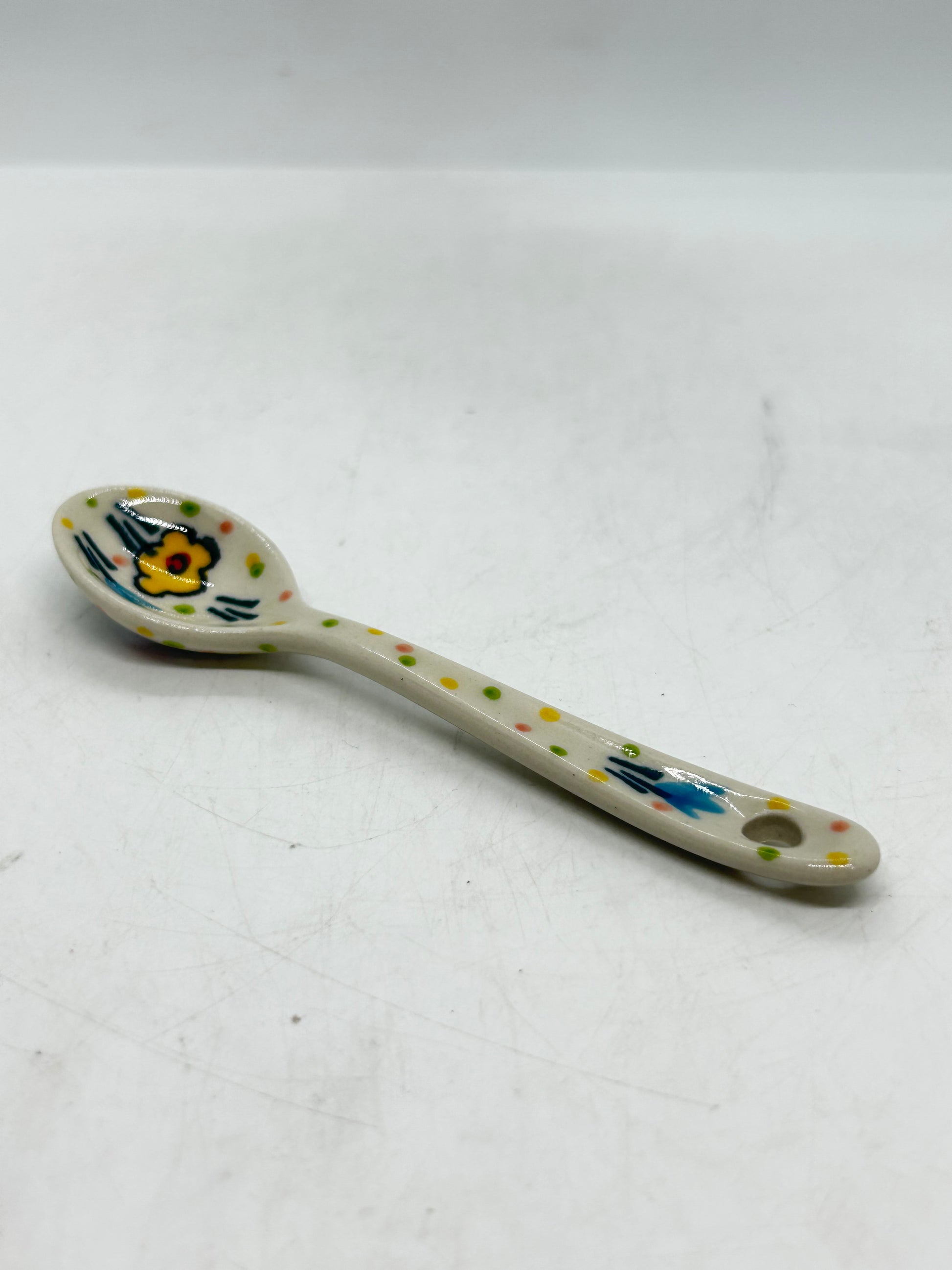 Elevated angled view of a small Ceramika Artystyczna Polish pottery spoon, featuring a hanging hole on the handle and a hand-painted yellow flower inside the basin.