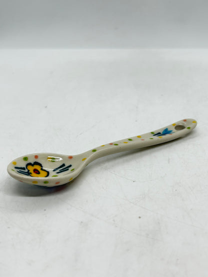 Ground-level side profile of a small Ceramika Artystyczna Polish pottery spoon resting on a white surface, showing an arched handle with a hanging hole, dotted pattern, and a hand-painted yellow flower inside the bowl.