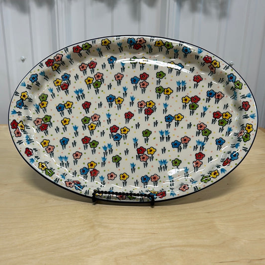 Straight-on display view of a large Ceramika Artystyczna Polish pottery oval serving platter on a stand, showing the full hand-painted spring floral pattern and dark blue rim.