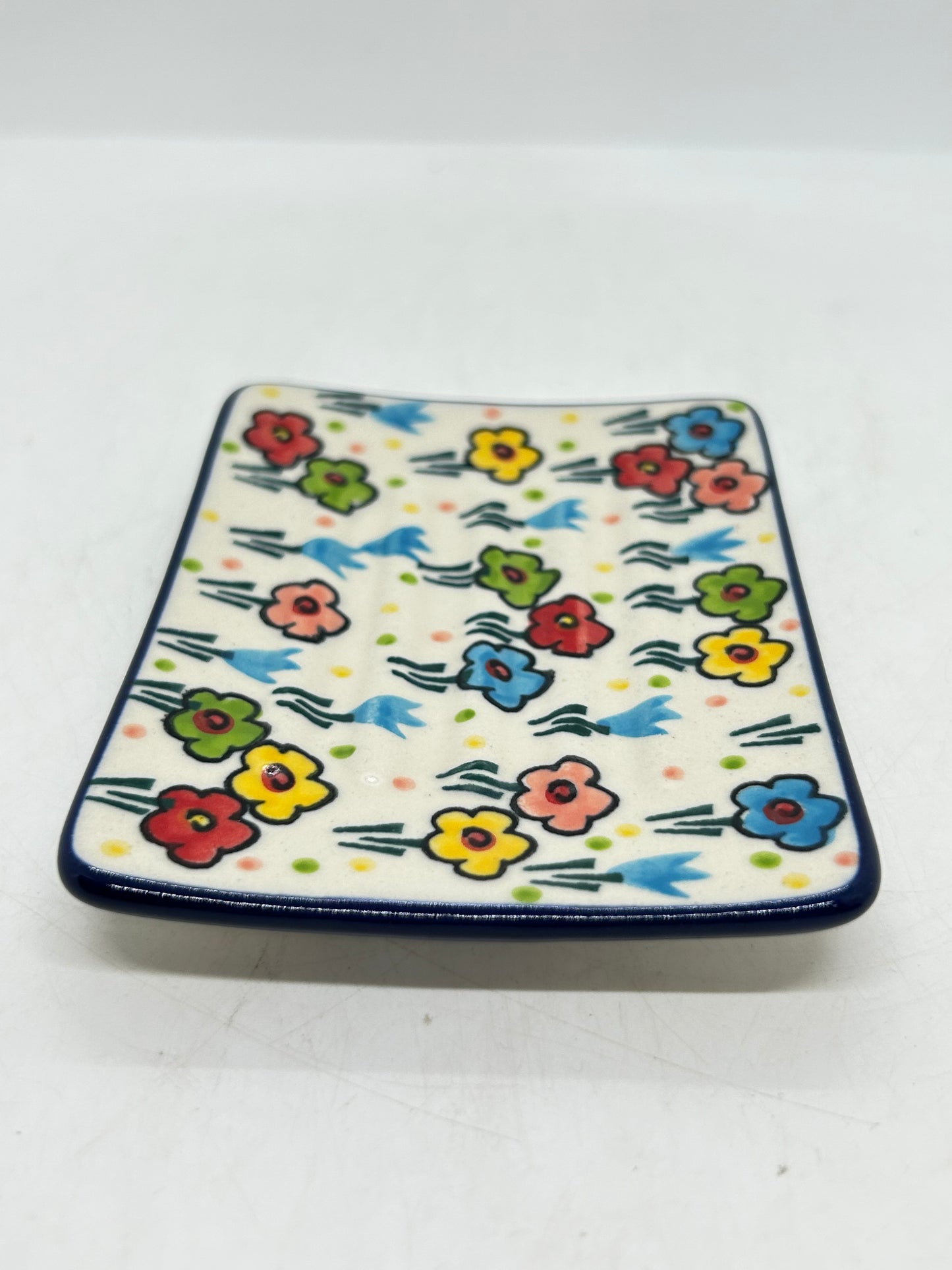 Angled view of a Ceramika Artystyczna Polish pottery rectangular soap dish showing ridged contours, a dark blue border, and hand-painted spring flowers.