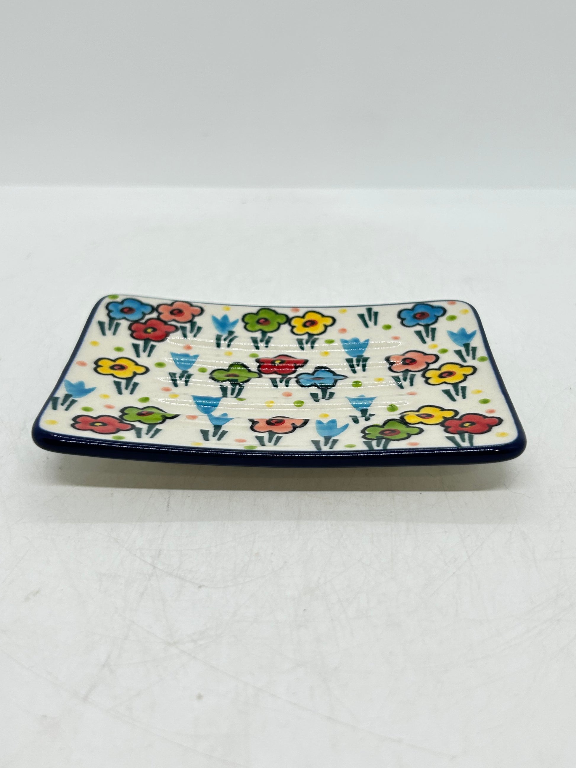 Low-angle side profile of a Ceramika Artystyczna Polish pottery rectangular soap dish showing ridged contours, a dark blue rim, and hand-painted spring flowers.