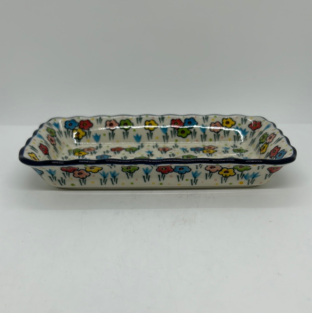 Ground-level side profile of a Ceramika Artystyczna Polish pottery rectangular serving tray, showing a low-profile silhouette, wavy dark blue rim, and hand-painted spring flowers wrapping around the exterior.