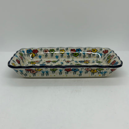 Ground-level side profile of a Ceramika Artystyczna Polish pottery rectangular serving tray, showing a low-profile silhouette, wavy dark blue rim, and hand-painted spring flowers wrapping around the exterior.