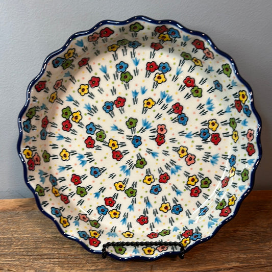 Straight-on view of a Ceramika Artystyczna Polish pottery fluted tart or quiche dish resting on a black wire stand, featuring a wavy dark blue rim and a fully decorated interior of hand-painted multi-colored spring flowers.