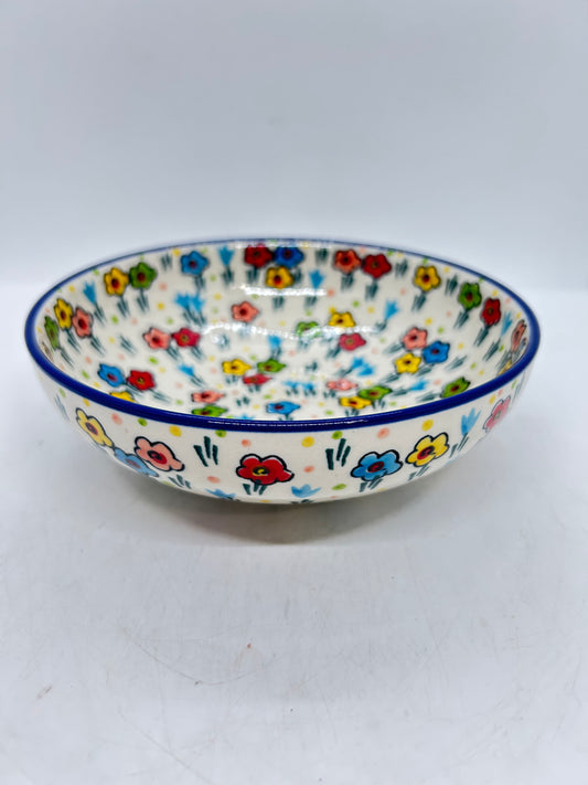 Slightly elevated angled view of a Ceramika Artystyczna Polish pottery shallow bowl, showing a dark blue rim and hand-painted multi-colored spring flowers covering the interior basin and exterior walls.