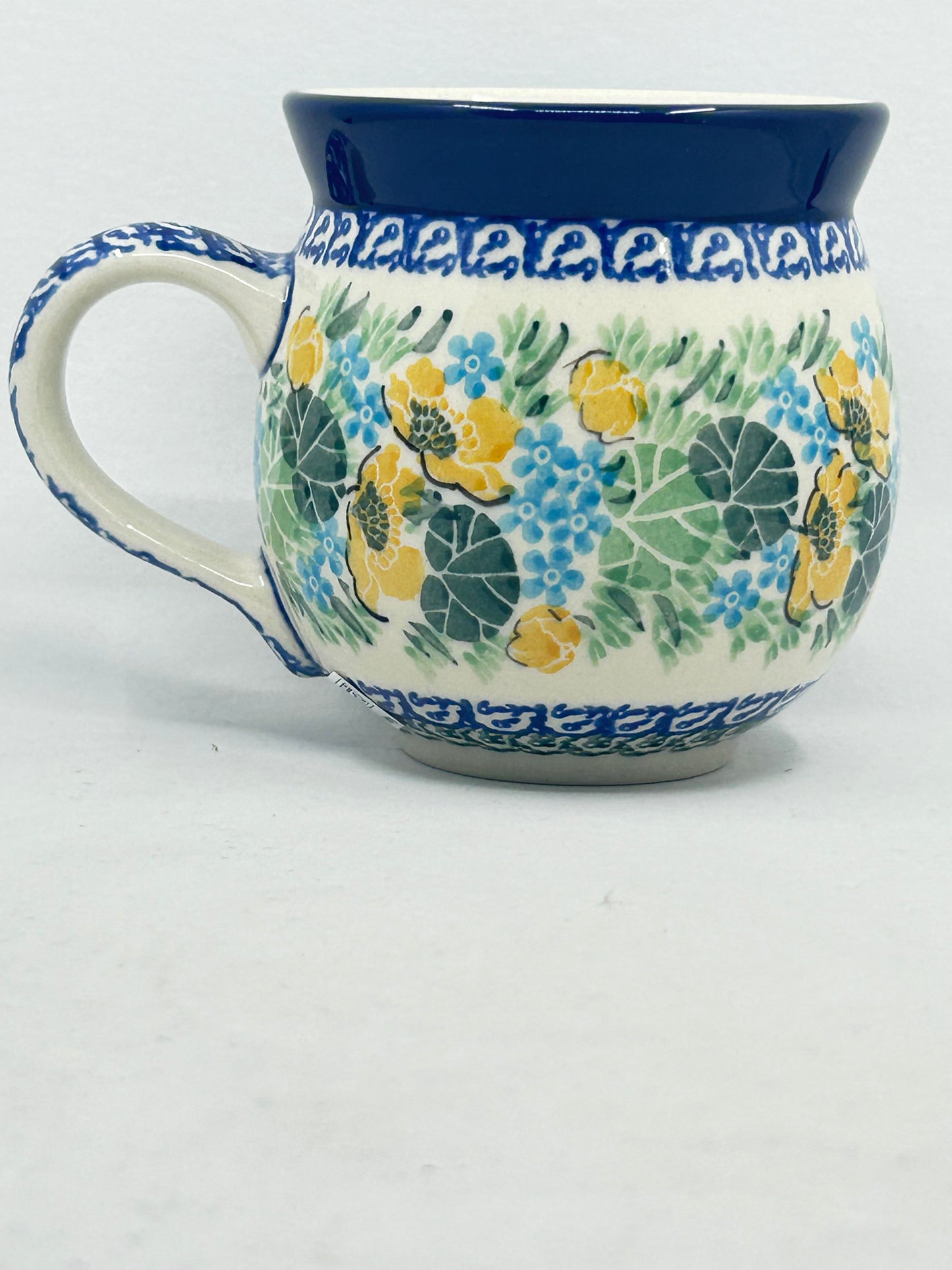 Straight-on side profile of a Ceramika Artystyczna Polish pottery bubble mug with the handle on the left, featuring hand-painted yellow and blue flowers, lush green leaves, patterned borders, and a solid dark blue rim.
