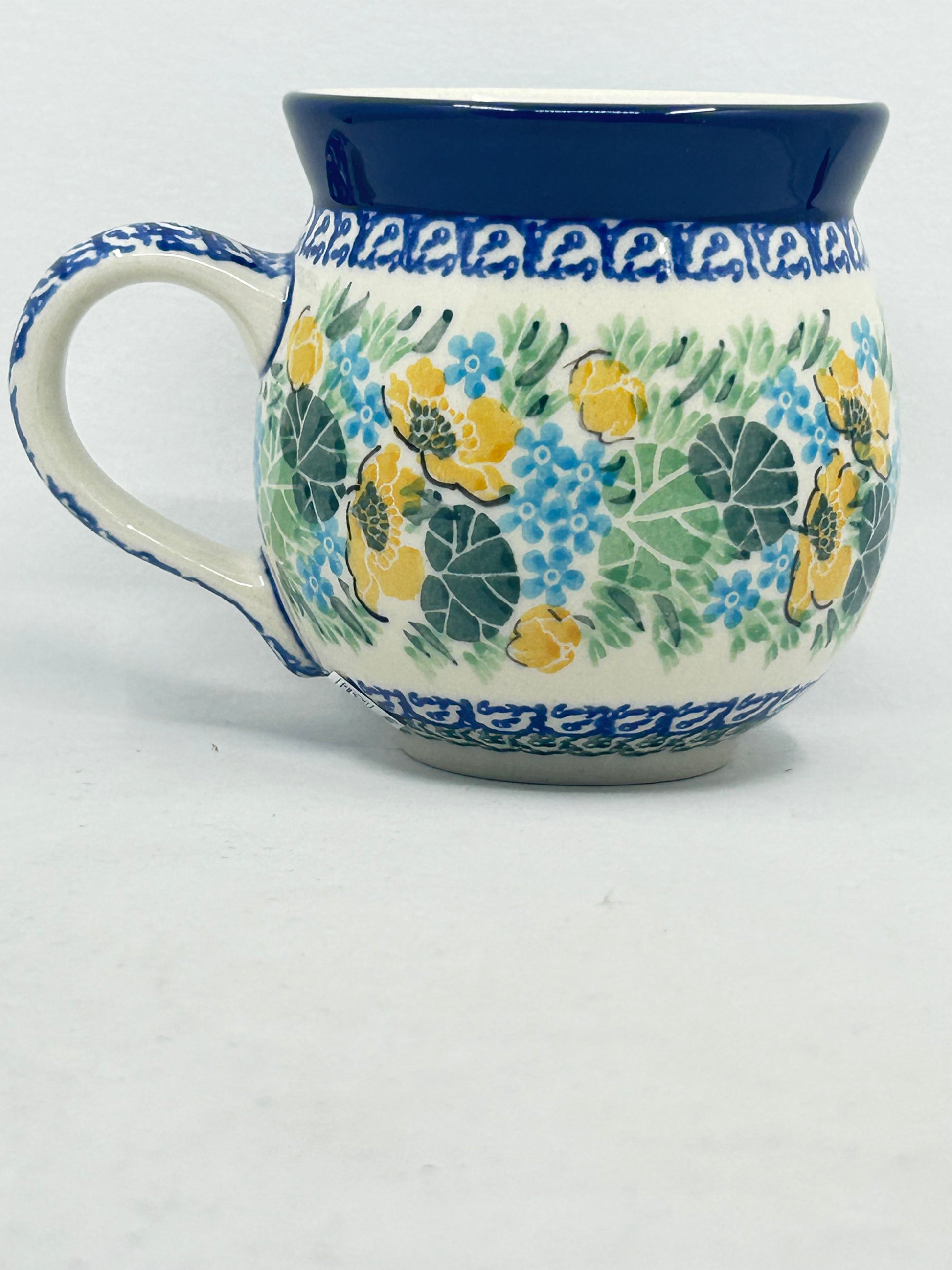 Straight-on side profile of a Ceramika Artystyczna Polish pottery bubble mug with the handle on the left, featuring hand-painted yellow and blue flowers, lush green leaves, patterned borders, and a solid dark blue rim.