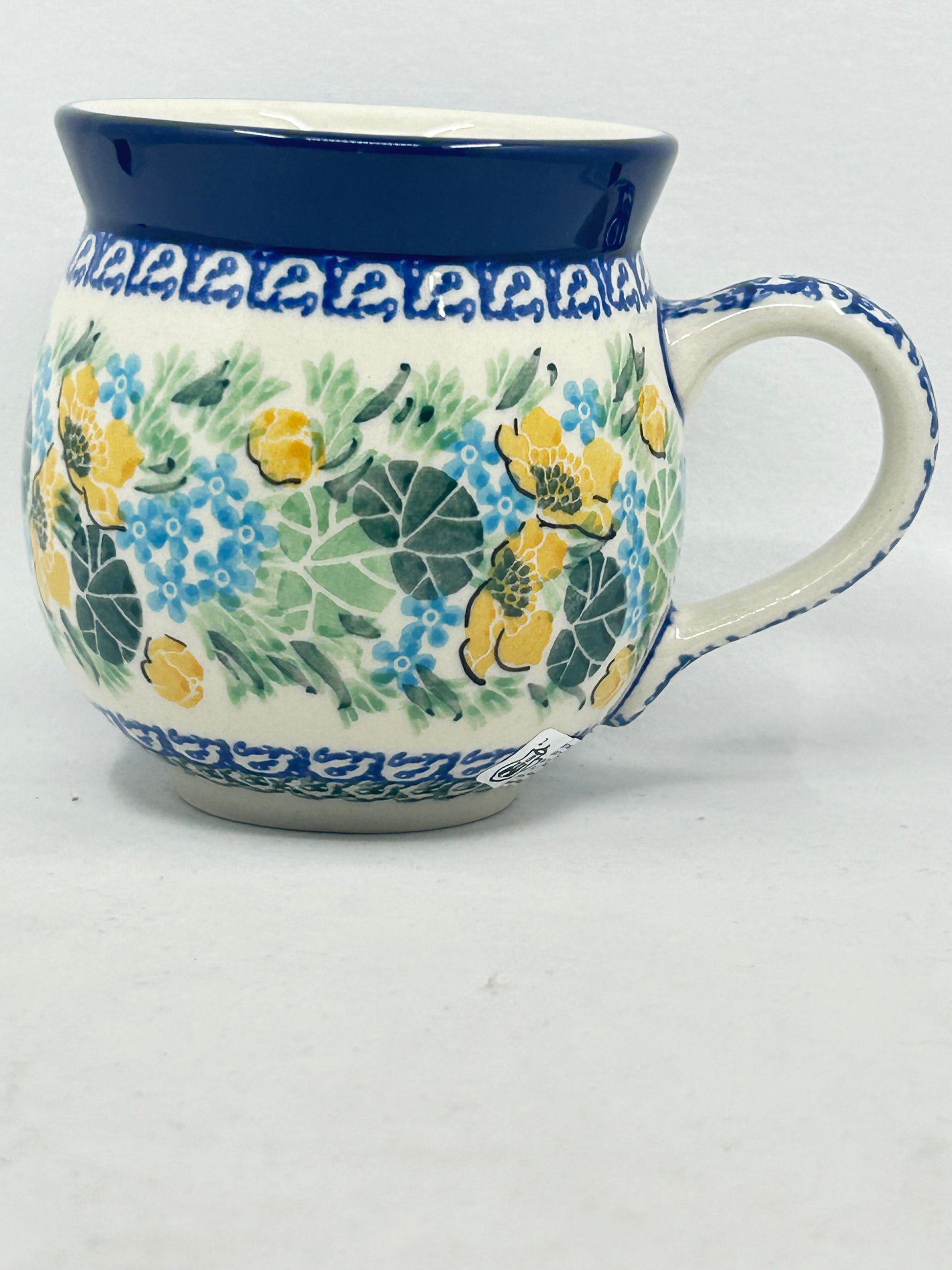 Straight-on side profile of a Ceramika Artystyczna Polish pottery bubble mug with the handle on the right, featuring hand-painted yellow and blue flowers, lush green leaves, patterned borders, and a solid dark blue rim.