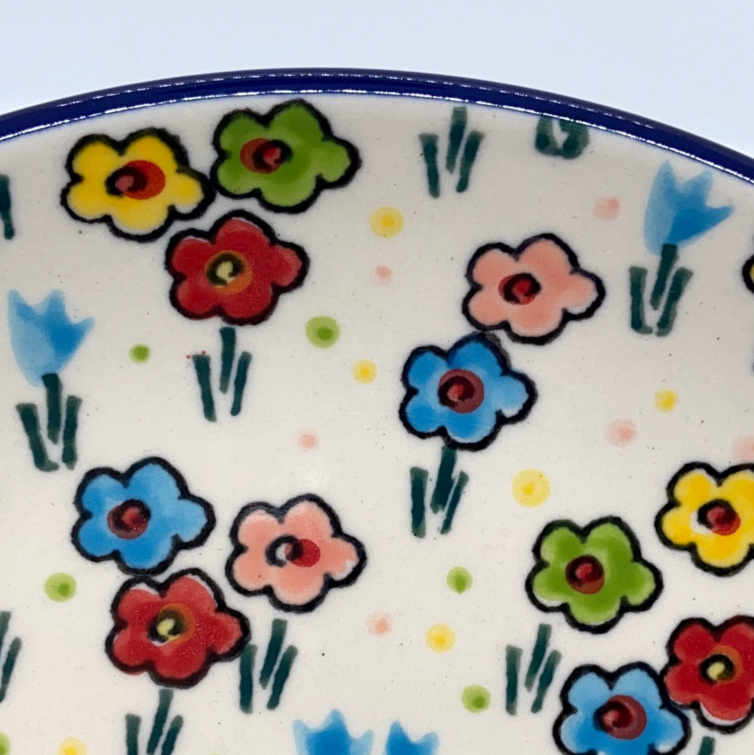 Close-up detail of a Ceramika Artystyczna Polish pottery plate, showing hand-painted yellow, red, blue, green, and pink flowers with dark outlines near a thin blue rim.