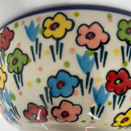 Extreme close-up of the curved exterior wall of a Ceramika Artystyczna Polish pottery bowl, showing a dark blue rim and distinct artisan brushstrokes on hand-painted multi-colored spring flowers.