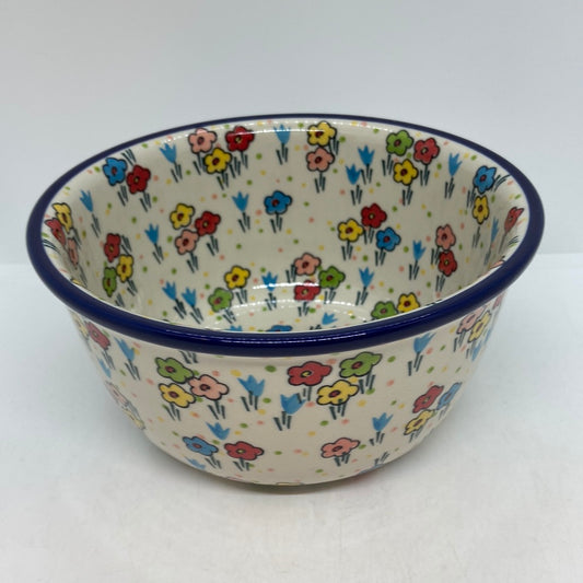 Slightly elevated angled view of a Ceramika Artystyczna Polish pottery deep round bowl, showing a dark blue rim and hand-painted multi-colored spring flowers covering the interior basin and exterior walls.